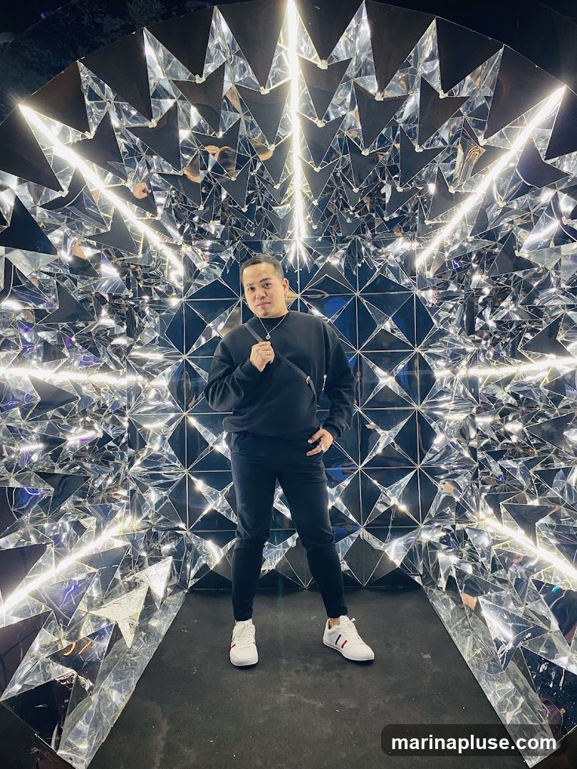 A man stands in the center of a room with walls covered in mirrored geometric shapes and illuminated by bright white lights. He is wearing a black sweatshirt, black pants, and white sneakers with red and blue stripes. The mirrored surfaces create a kaleidoscopic effect, reflecting the lights and the man's image multiple times.