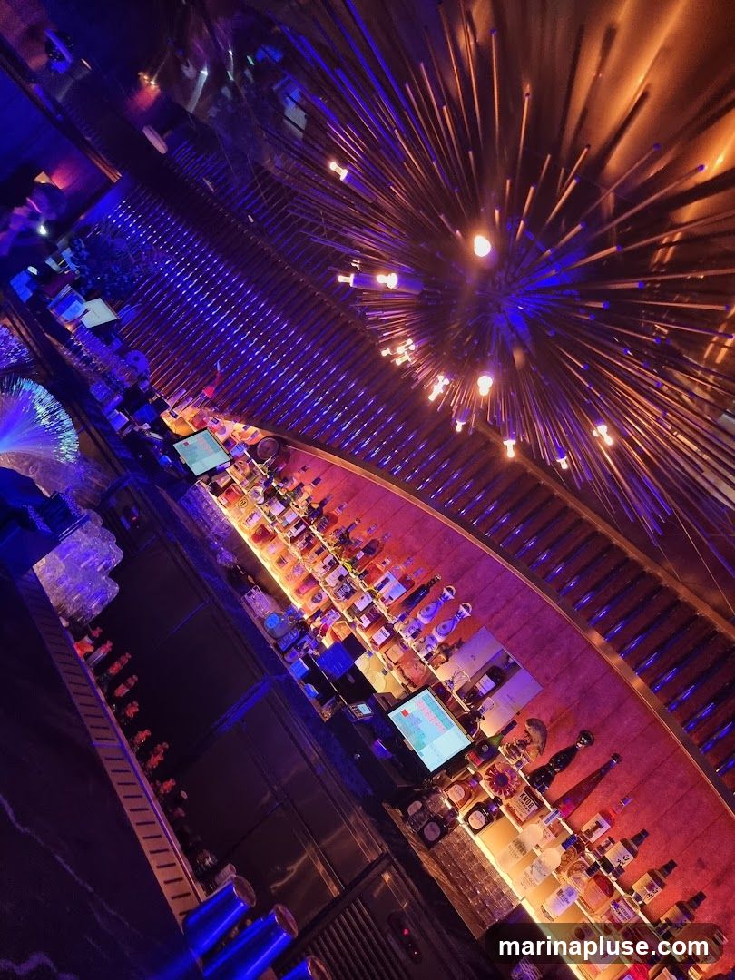 An overhead view of a bar interior with bottles of alcohol displayed on shelves behind the bar. There are also glasses, a computer monitor, and a unique light fixture on the ceiling. The lighting is dim and colorful, with blue and orange hues.