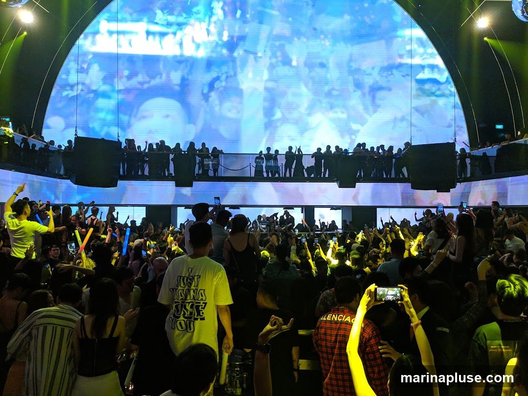 A large crowd of people are gathered in a dimly lit venue, with many holding up their phones and glow sticks. A large screen displays abstract, colorful imagery. Silhouettes of people can be seen on an upper level overlooking the main crowd. The atmosphere appears energetic and celebratory.