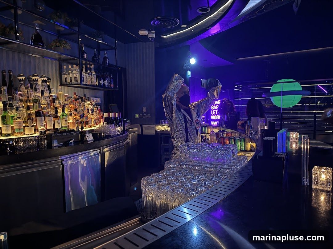 A dimly lit bar interior with a long counter stocked with numerous bottles of alcohol. A bartender in a silver, futuristic-looking outfit stands behind the counter, wearing a black face mask. Neon signs are visible in the background, including one that reads "COME BAP LET'S MARRY". A green, planet-like decoration hangs from the ceiling. Stacks of clean glasses are arranged on the counter.
