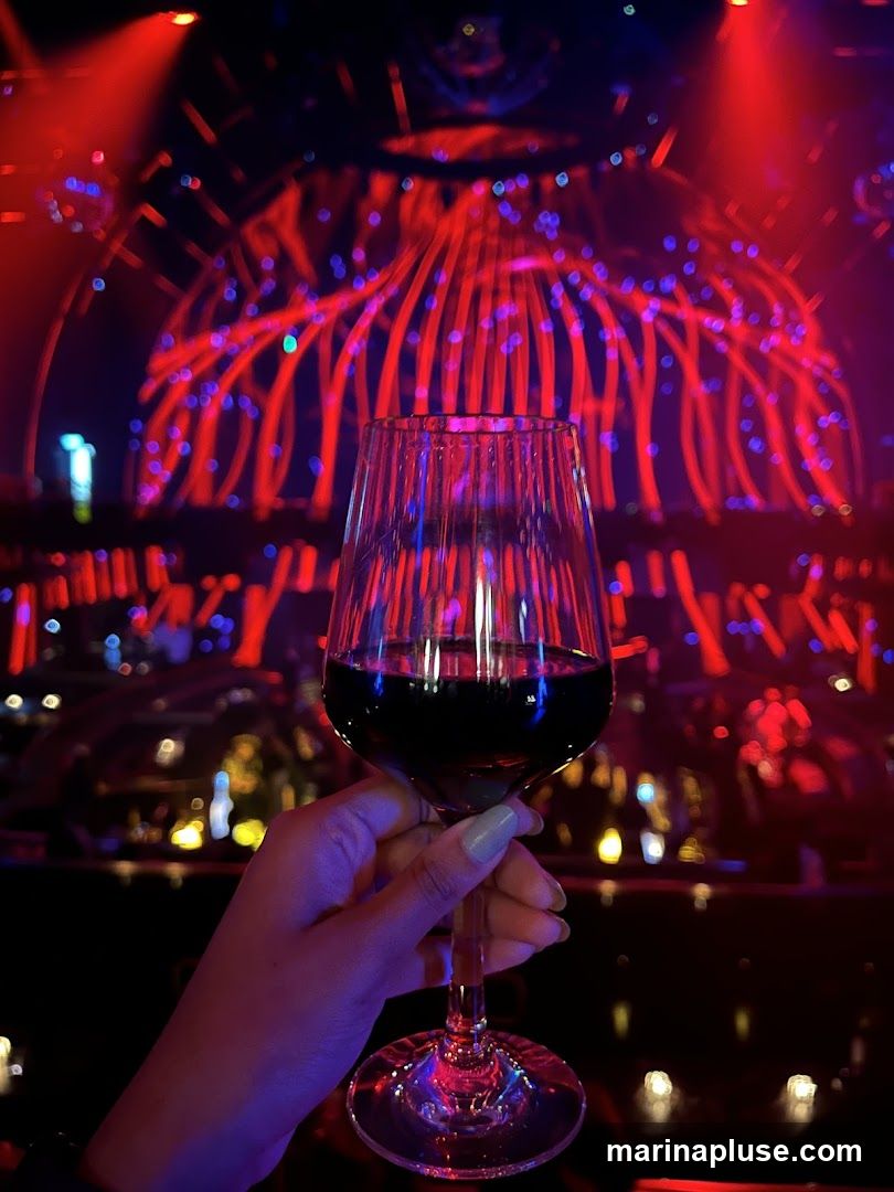 A hand holds a glass of red wine in the foreground, with a blurred background of a nightclub or concert venue. The background features dramatic red and blue lighting, with illuminated structures and bokeh effects from lights.