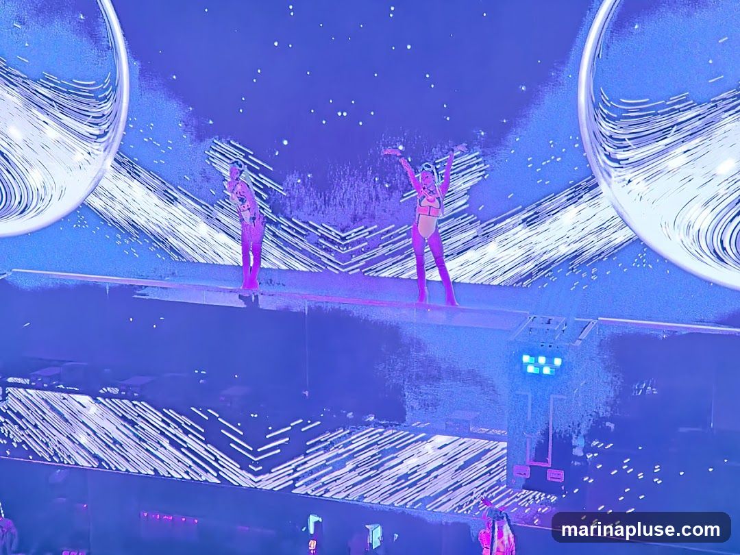 Two performers in bright outfits are on a stage with a large, abstract, blue and white background. The stage is lit with blue lights, and there are large, spherical elements on either side of the background.