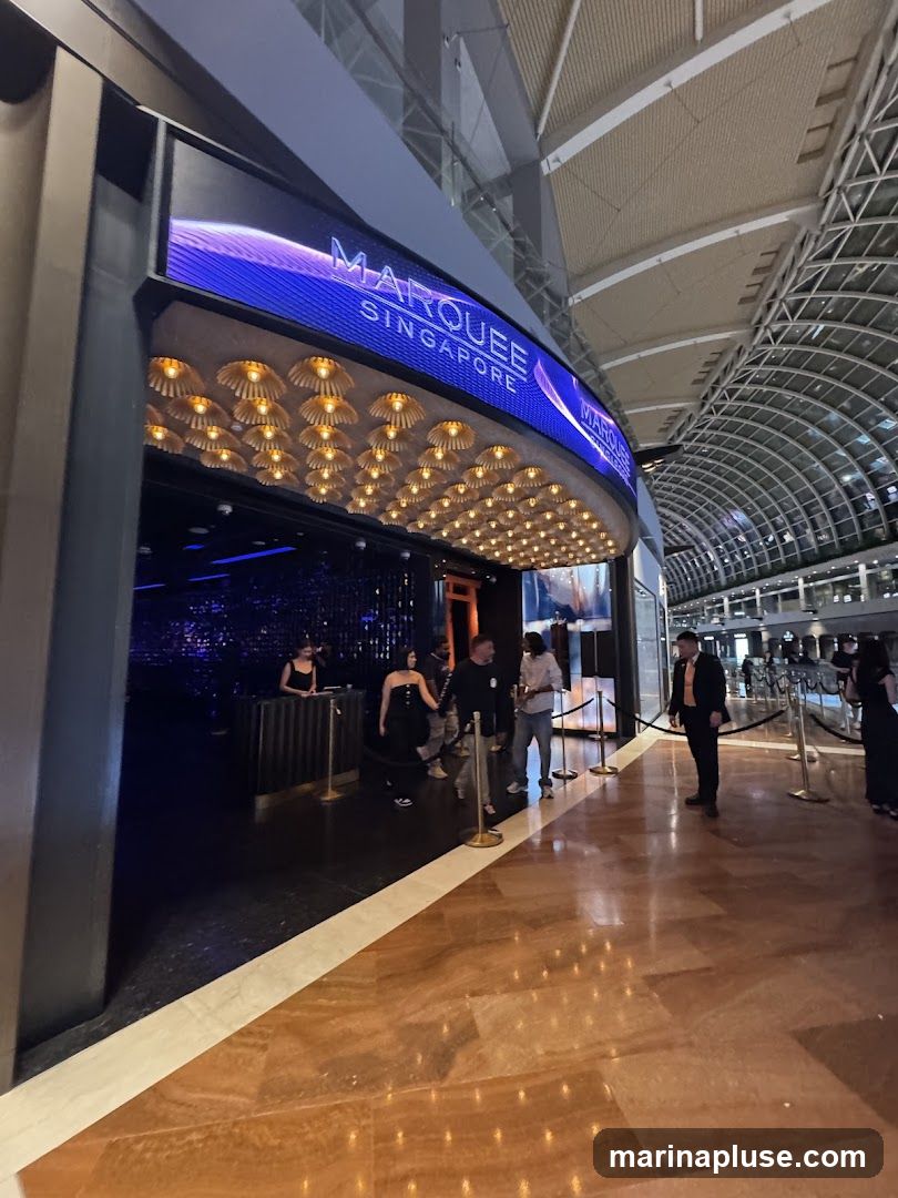 The entrance to Marquee Singapore is illuminated by a large LED screen displaying the club's name and a wavy purple design. Below the screen, a row of decorative golden lights adds to the ambiance. People are seen entering the venue, with staff members managing the queue. The floor is made of polished marble, reflecting the lights and creating a luxurious feel.