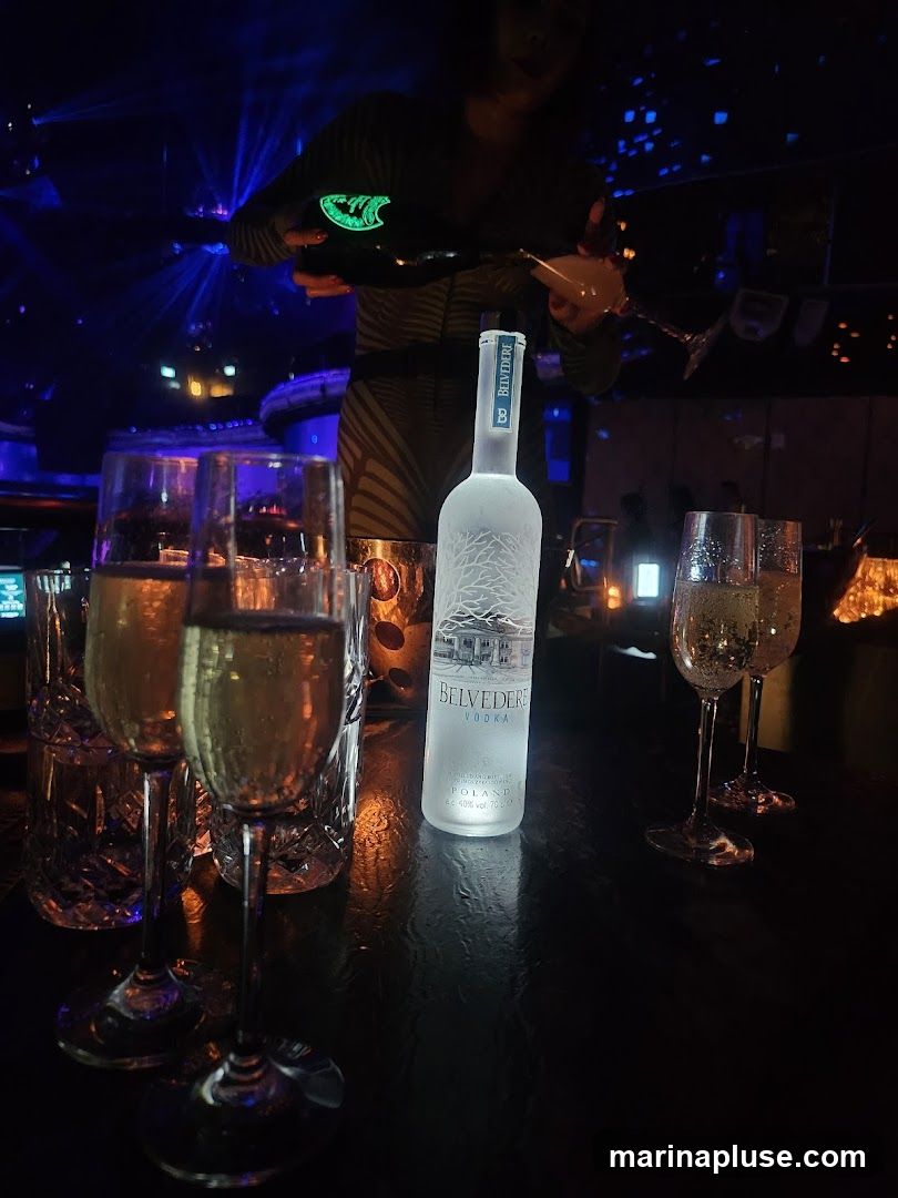 A person is pouring champagne into a glass from a bottle. A bottle of Belvedere vodka stands on a dark, reflective surface, surrounded by several champagne flutes filled with bubbly liquid. The background is dimly lit with blue and purple lights, suggesting a nightclub or bar atmosphere.