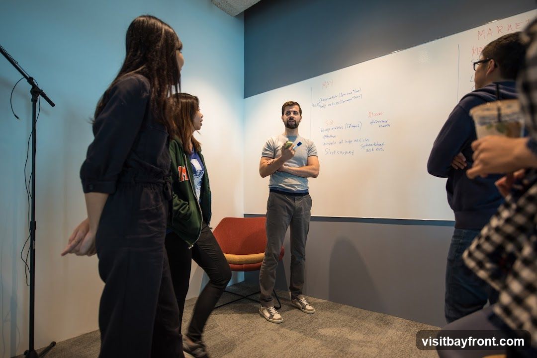 A group of people are gathered in a room, with one man standing in front of a whiteboard. The whiteboard has handwritten notes and tasks listed under headings like "MAY" and "MARKET". The man is holding a marker and appears to be leading a discussion. The other individuals are observing the whiteboard or listening to the speaker.