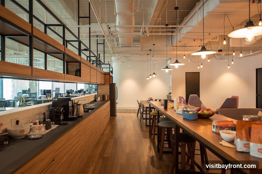A contemporary office break room with a long wooden counter featuring a coffee machine, cups, and bowls. Behind the counter, a glass partition reveals an open-plan office space with desks and computers. The room also includes a communal dining table with stools, chairs, and a fruit basket. Industrial-style pendant lights hang from the exposed ceiling.