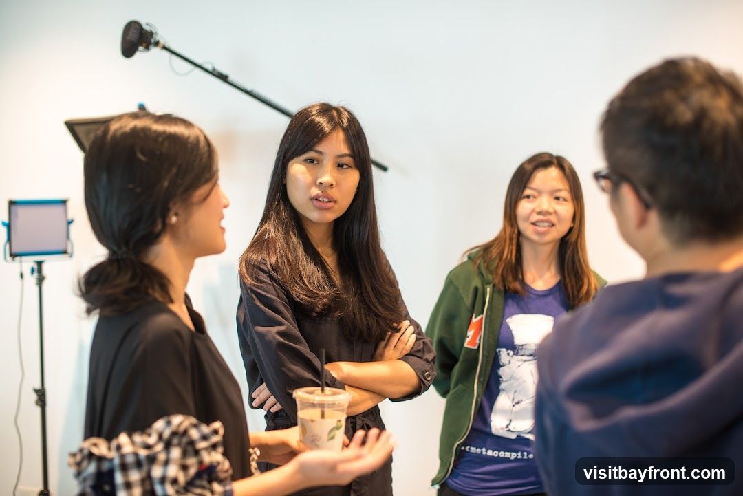 Four people are gathered in a room, possibly for a meeting or a discussion. One person in the foreground is holding a drink. In the background, there is a microphone on a boom pole and a lighting setup, suggesting a video recording or presentation is taking place. The people are casually dressed.