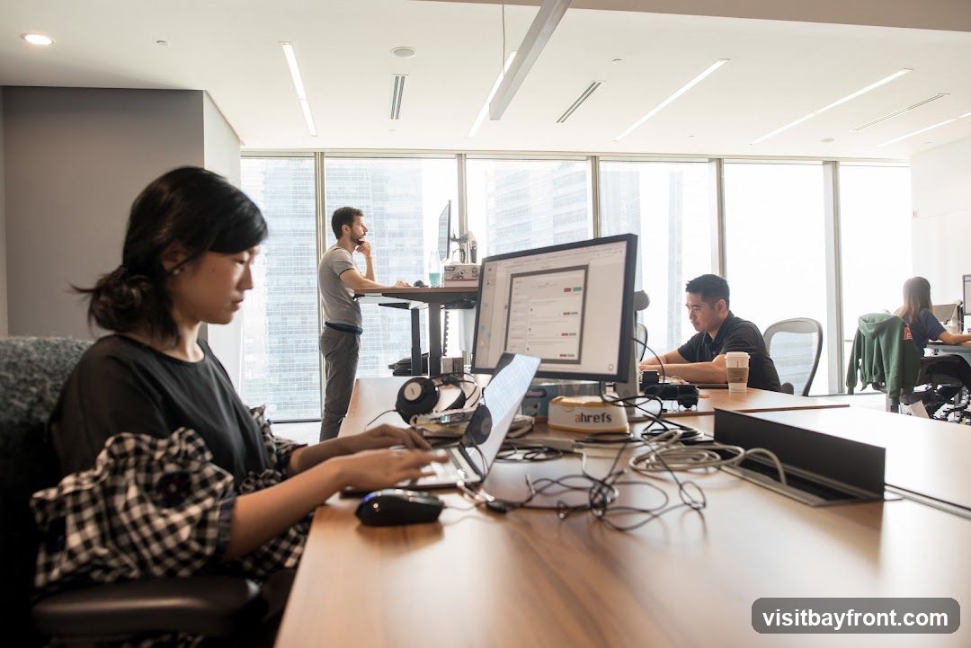 A modern office interior with several people working at desks. In the foreground, a woman is typing on a laptop. To her right, a man is working at a computer, and a coffee cup is on the desk. In the background, a man stands at a standing desk, and another person is visible at a desk further away. Large windows offer a view of city buildings.