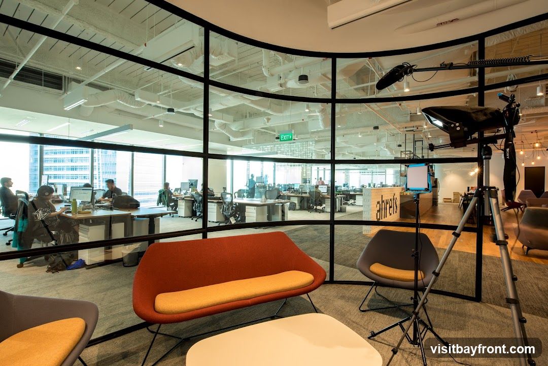 A view into a modern office space through a curved glass wall. The office features open-plan workstations with employees working at computers. In the foreground, there are comfortable seating areas with modern chairs and a coffee table. A video recording setup, including a camera, lights, and microphone on a tripod, is positioned in the foreground, suggesting a filming or content creation activity within the office. The company name 'ahrefs' is visible on a reception desk or partition inside the office.