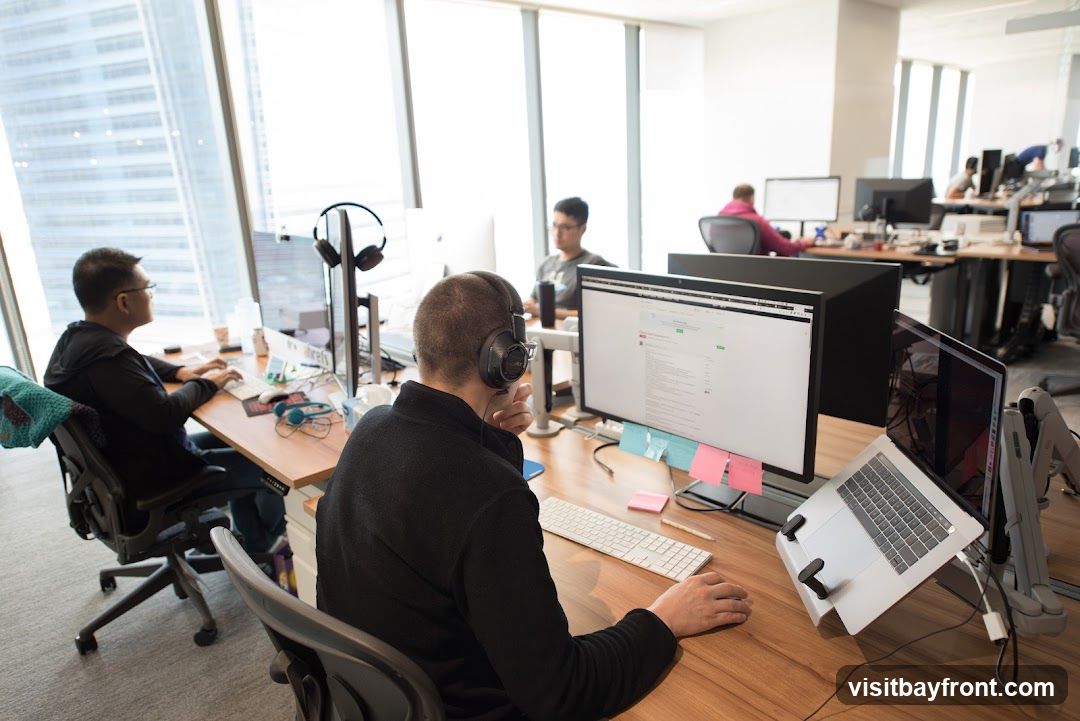 Several people are working at computers in a modern office. The desks are made of wood and equipped with multiple monitors, keyboards, and laptops. Some individuals are wearing headphones. The office has large windows with a view of city buildings.