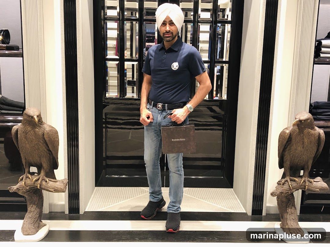 A man wearing a blue polo shirt, jeans, and a white turban stands in a store. He is holding a brown shopping bag with the logo "STEFANO RICCI" visible. The store appears to be a high-end boutique, with displays of clothing and decorative eagle statues on either side of the entrance. The man is standing in front of a large, reflective door with black and white trim.