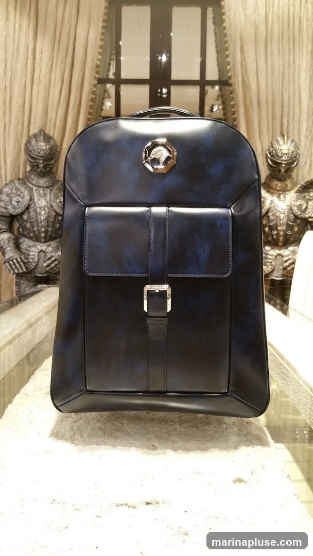 A black leather backpack with a silver buckle and a metallic emblem on the front is displayed. The backpack has a front pocket with a flap and a strap closure. It is placed on a textured surface, possibly stone, with two suits of armor visible in the background, flanking a window with draped curtains.