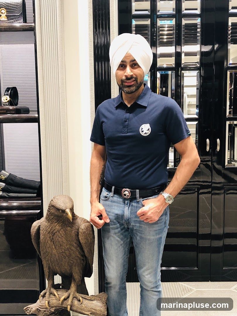 A man wearing a navy blue polo shirt, light blue jeans, and a white turban stands in a store. He is next to a large bronze statue of a hawk perched on a log. Behind him is a display case with various luxury items and a black, reflective door. The man has a watch on his left wrist and a belt with a distinctive buckle.
