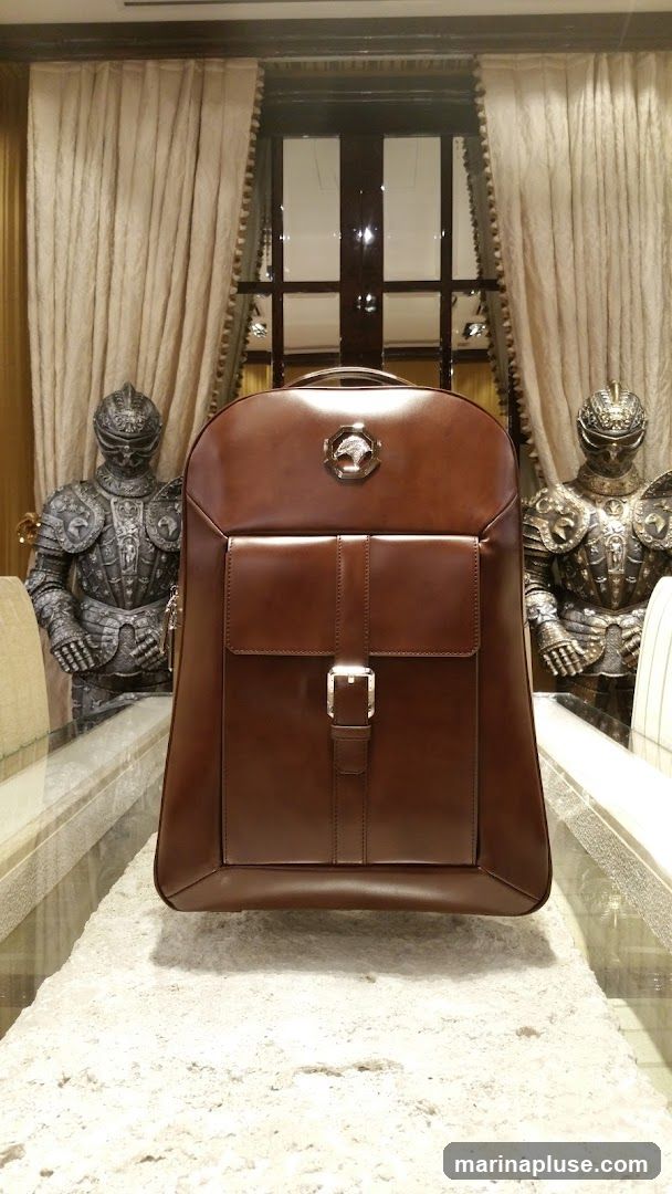 A brown leather backpack is displayed prominently in the center of the image. It features a large front pocket with a buckle closure and a metallic emblem at the top. The backpack is placed on a textured surface, possibly a table or display stand. Behind the backpack, two full suits of armor stand guard, flanking a large window with ornate curtains. The overall setting suggests a luxurious or themed retail environment.