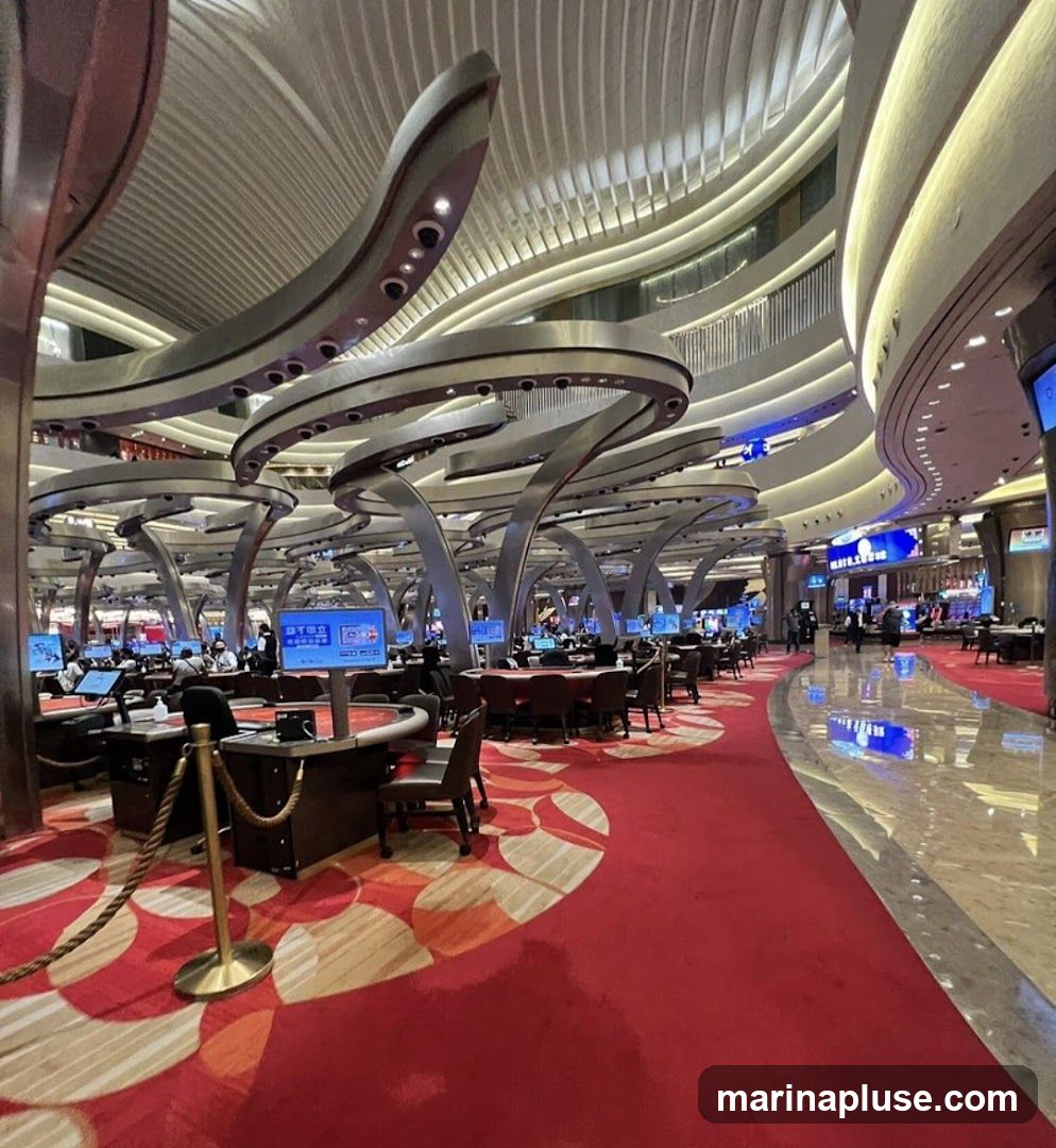 A wide shot of a casino gaming floor with numerous tables and electronic screens. The room features a modern, curved architectural design with metallic structures and bright lighting. Red carpets with a circular pattern cover the floor, and ropes guide pathways. Several people are seated at gaming tables, some interacting with dealers and others with the electronic displays.