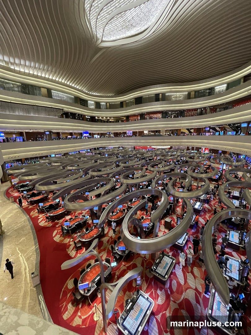 A high-angle view of a large, modern casino floor with numerous gaming tables and slot machines. The ceiling features a unique, undulating design with integrated lighting. The floor is covered in a red carpet with a vibrant, abstract pattern. People are seated at the tables, engaged in gaming.