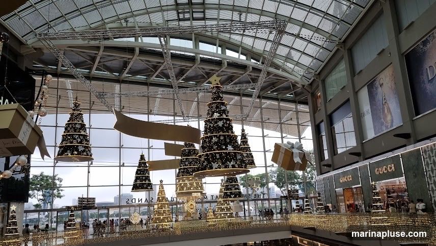 A wide shot of the interior of a shopping mall decorated for Christmas. Several large, tiered Christmas trees are suspended from the ceiling, adorned with lights and ornaments. Giant gift boxes are also hanging, adding to the festive atmosphere. The background shows storefronts of luxury brands like Dior and Gucci, with shoppers visible on the lower levels. The mall features a glass ceiling and large windows, allowing natural light to filter in.