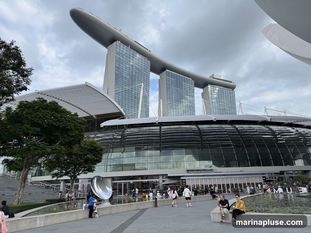 A wide shot of the Marina Bay Sands complex in Singapore on a cloudy day. The iconic three towers with the ship-like structure on top are visible in the background. In the foreground, the curved glass roof of the convention center is prominent, with people walking around the plaza and sitting by a water feature. A large, reflective sculpture is also present.