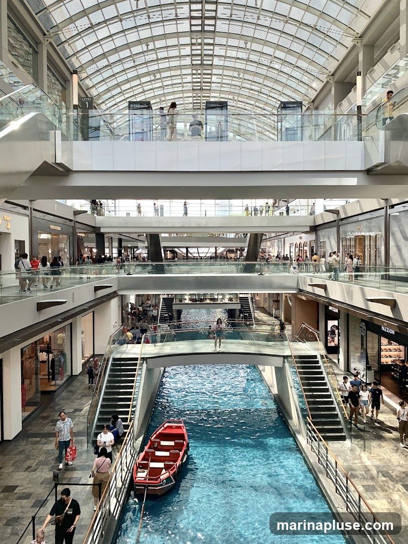 An interior view of a multi-level shopping mall with a canal running through the center. A small red boat is docked in the canal. People are walking on the walkways and bridges, and browsing in the shops. The mall has a large glass roof that allows natural light to enter.