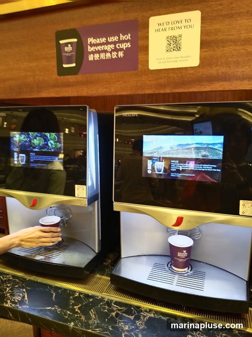 Two Nescafe beverage dispensers are shown. A hand is placing a cup under the left dispenser. The right dispenser has a cup with a lid. Signs above the dispensers read "Please use hot beverage cups" and "WE'D LOVE TO HEAR FROM YOU" with a QR code. The screens on the dispensers show images and text related to beverages.