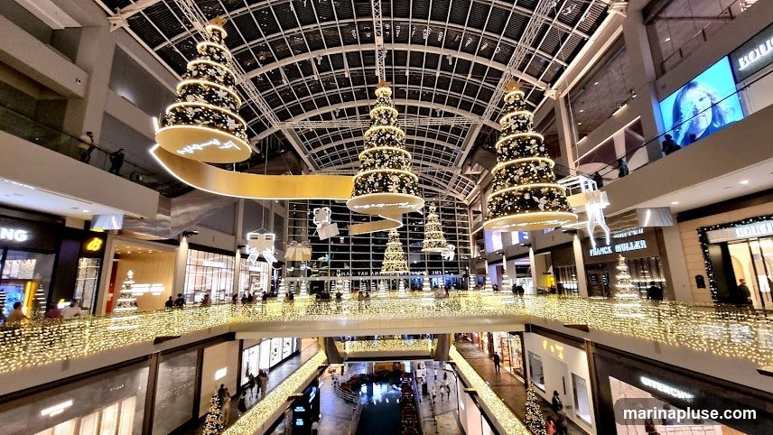 A wide shot of the interior of a multi-level shopping mall decorated for Christmas. Several large, tiered Christmas trees adorned with lights and ornaments hang from the ceiling. A golden ribbon-like structure with illuminated text winds through the space. Balconies and walkways are lined with strings of warm white lights. Various shops are visible on different levels, including 'BOUCHERON' and 'FRANCK MULLER'. People are seen walking around the mall.