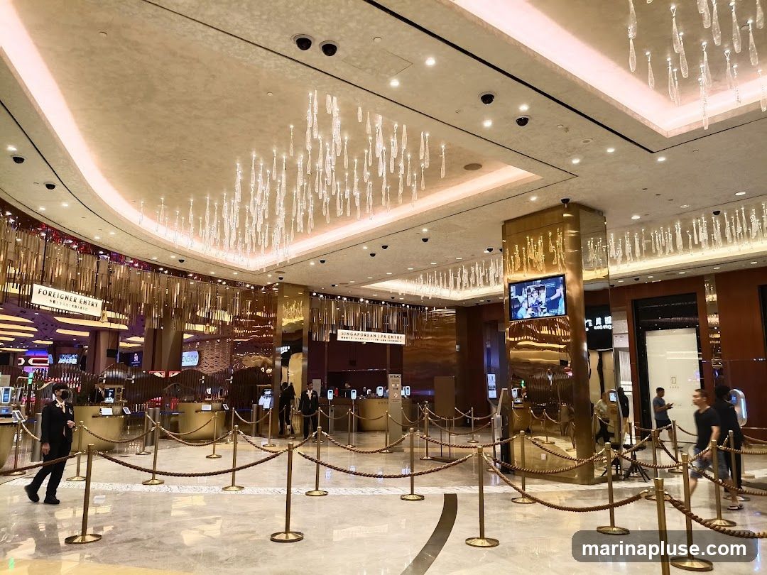 A wide shot of a luxurious casino lobby with ornate chandeliers and golden accents. There are multiple check-in counters with ropes guiding a queue. Signs indicate "FOREIGNER ENTRY" and "SINGAPOREAN ENTRY". People are seen walking through the lobby and at the counters. A large screen displays content on a golden pillar.