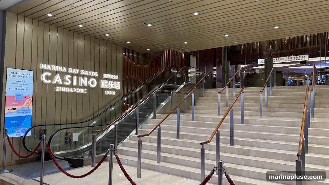 The image shows the entrance to the Marina Bay Sands Casino in Singapore. The main entrance is currently closed, and a sign directs visitors to an alternative entrance on Level B1. The signage for "MARINA BAY SANDS CASINO SINGAPORE" is prominently displayed. Escalators and stairs lead up to the casino entrance.