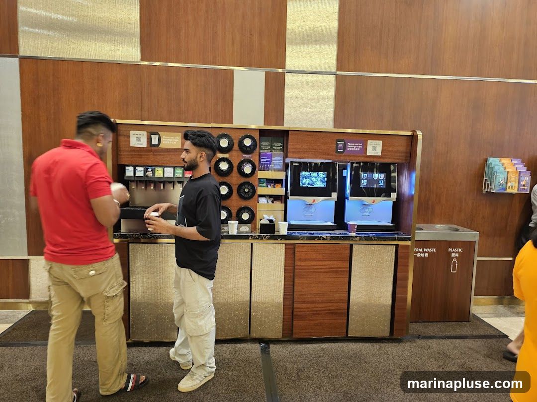 Two men are standing at a beverage station with multiple drink dispensers and coffee machines. One man in a red shirt is holding a cup, and the other man in a black t-shirt is also holding a cup. There are screens on the coffee machines displaying graphics. To the right, there is a recycling bin labeled "ERAL WASTE" and "PLASTIC". A rack of brochures is mounted on the wall above the bin.