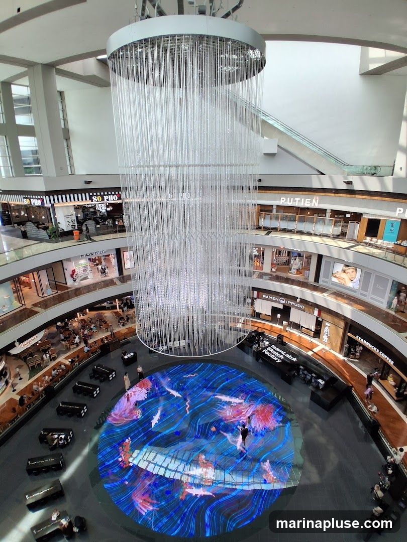 An overhead view of a multi-level shopping mall atrium. A large, modern chandelier hangs from the ceiling, with numerous clear strands descending towards a circular digital display on the floor. The display shows a vibrant, animated scene with koi fish and coral-like structures. Surrounding the atrium are various shops and escalators. Visible shop signs include "SO PHO", "FILA KIDS", "PUTIEN", "RAMEN IPPUDO", "teamLab Sketch Factory", and "STARBUCKS COFFEE". People are seen browsing shops and walking around the mall.