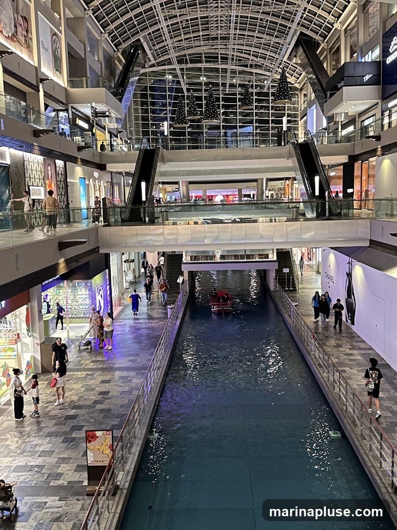 An interior view of a multi-level shopping mall with a canal running through the center. Escalators and walkways connect the different levels, and various shops are visible along the sides. A small boat navigates the canal. Christmas trees are suspended from the ceiling.
