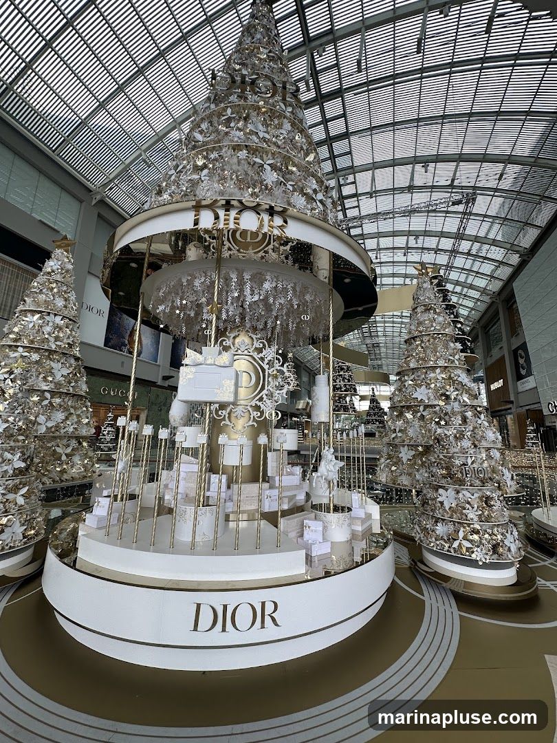 A large, ornate Christmas tree display with the Dior logo prominently featured. The display is decorated with white and gold ornaments and features smaller Christmas trees around it. The background shows a shopping mall interior with other store signs visible.