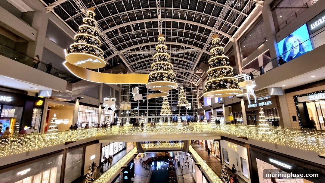 A wide shot of the interior of a multi-level shopping mall decorated for Christmas. Large, tiered Christmas trees adorned with lights and ornaments hang from the ceiling. Balconies and railings are draped with strings of warm white lights. Shops with illuminated signage are visible on different levels, including Franck Muller and Boucheron. People are seen walking on the escalators and walkways. A large digital screen displays a woman's face.
