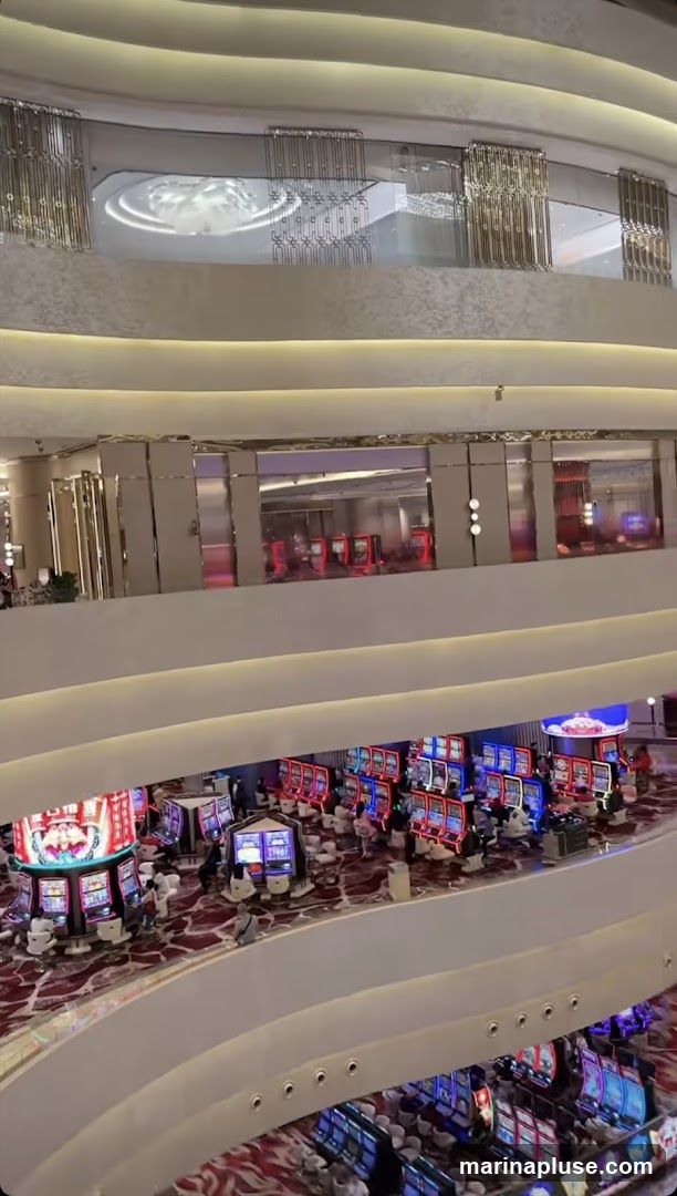 A multi-level casino interior with numerous slot machines and patrons. The decor is modern and luxurious, with curved balconies, ornate lighting fixtures, and plush carpeting.