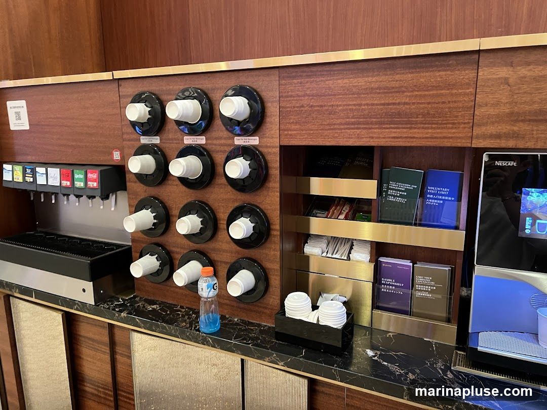 A beverage station with a soda fountain, cup dispensers, tea and coffee supplies, and a Nescafé machine. The soda fountain has dispensers for various sodas including Coke, Sprite, and Fanta. There are multiple cup dispensers with white paper cups. To the right, there are shelves with tea bags, sugar packets, and other amenities. A Nescafé coffee machine is visible on the far right. Several informational pamphlets are also displayed.