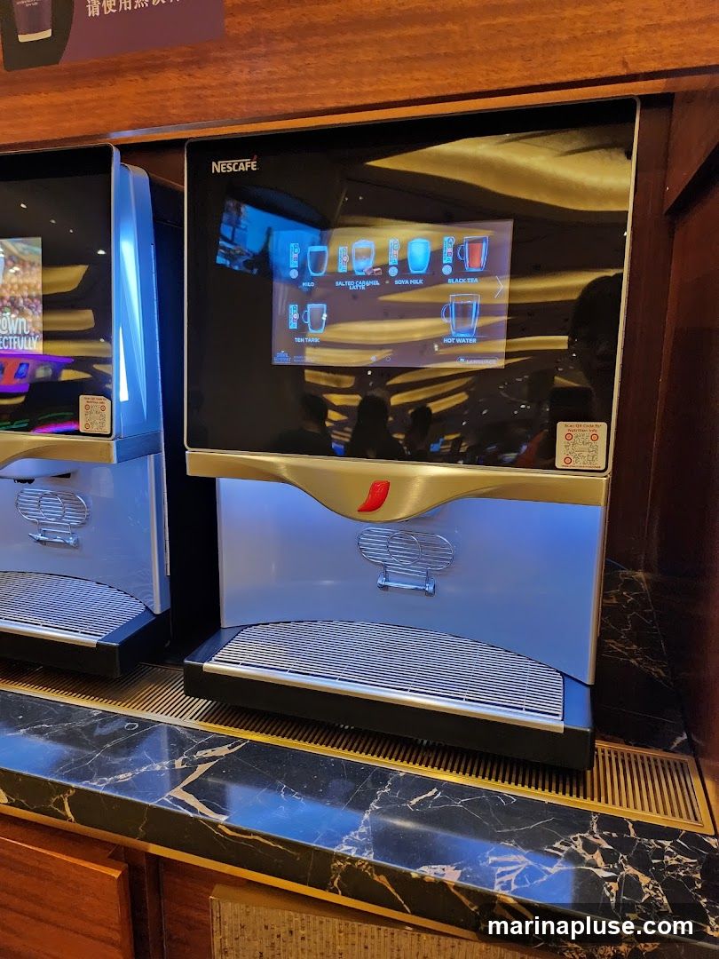 A Nescafé branded drink dispenser with a touchscreen interface displaying various beverage options. The screen shows icons for Hilo, Salted Caramel, Soya Milk, Black Tea, Teh Tarik, and Hot Water. The dispenser is situated on a marble countertop with a metallic grate.