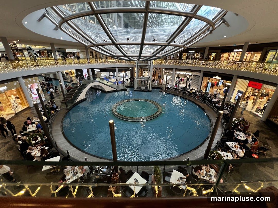 An interior view of a multi-level shopping mall featuring a large circular indoor canal or water feature with a fountain in the center. Shops line the upper levels, some with visible signage like HUGO. Balconies overlook the water feature, and tables and chairs are set up around the perimeter for dining. Decorative lights are strung along the railings.