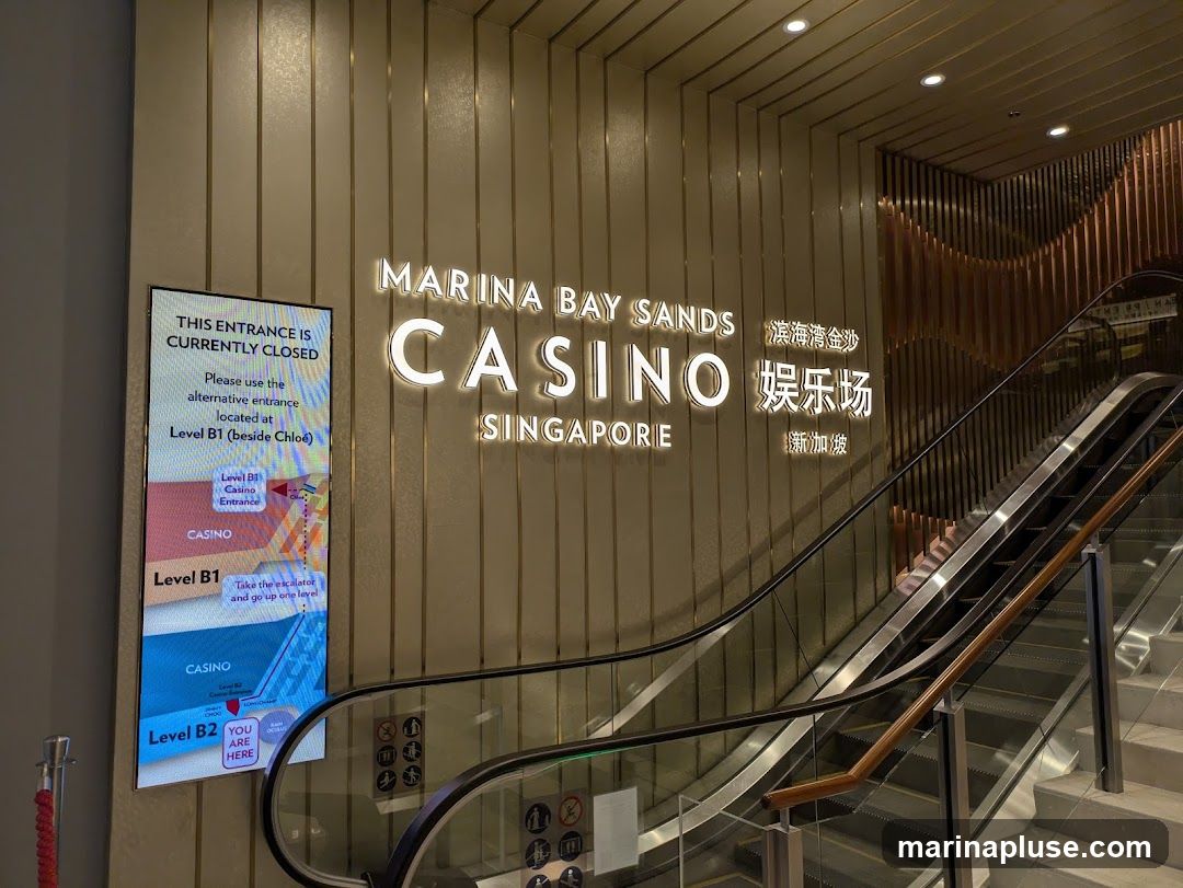 A digital sign displays that the current entrance is closed and directs visitors to an alternative entrance at Level B1, beside Chloé. The sign also includes a map showing the layout of the casino levels. Above the sign, illuminated text reads "MARINA BAY SANDS CASINO SINGAPORE" in English and Chinese. An escalator is visible to the right of the sign.