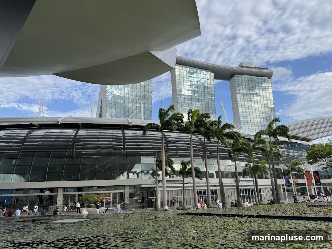 A wide shot of the Marina Bay Sands complex in Singapore, featuring its iconic three towers and the SkyPark. In the foreground, a body of water with lily pads is visible, and palm trees line the walkway in front of the building's glass facade. People are seen walking and sitting by the water. The sky is partly cloudy.