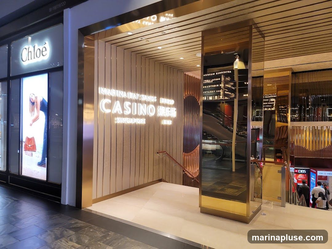 The entrance to the Marina Bay Sands Casino in Singapore is shown. The entrance features a modern design with vertical wooden panels and gold accents. The words "MARINA BAY SANDS CASINO SINGAPORE" are illuminated in white lettering. To the left, a Chloe store is visible with a lit advertisement. To the right, escalators and signs for various attractions, including a museum and nightclub, are reflected in mirrored surfaces.