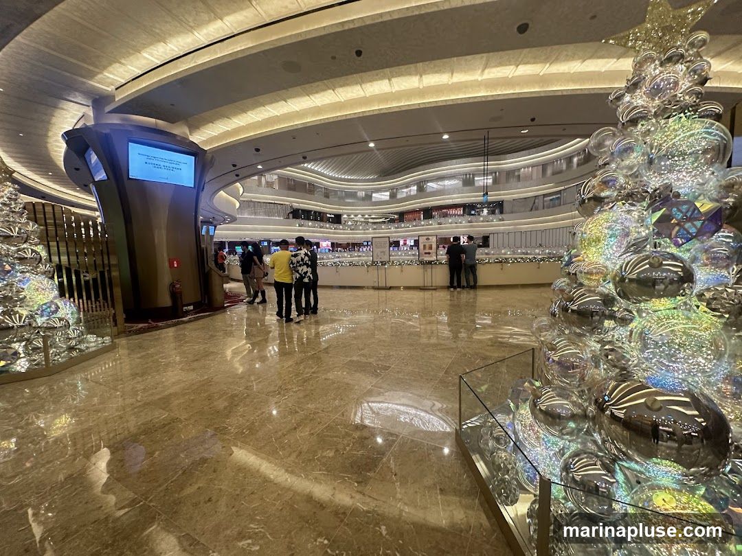A wide shot of a modern, multi-level interior space, possibly a shopping mall or casino, decorated for the holidays. In the foreground, a large, shimmering Christmas tree made of reflective ornaments and geometric shapes stands prominently. The floor is polished marble, reflecting the ambient light from the curved ceiling and the various levels of the building visible in the background. Several people are scattered throughout the space, some gathered near a display screen on a curved pillar, others looking at displays on a railing in the mid-ground. The overall impression is one of a spacious, upscale, and festive environment.