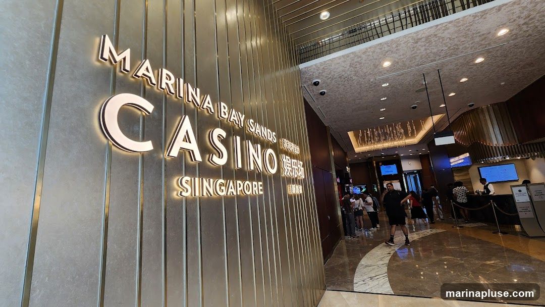 A large, illuminated sign reads "MARINA BAY SANDS CASINO SINGAPORE" in prominent white lettering against a textured metallic wall. The entrance to the casino is visible in the background, with people queuing and staff at a reception desk. The interior features a grand ceiling with decorative lighting and a polished marble floor.