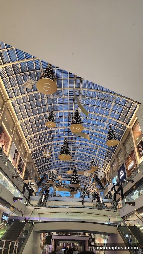 The image shows the interior of a multi-level shopping mall with a glass ceiling. Several Christmas trees decorated with stars and Dior logos are suspended from the ceiling. Escalators and walkways are visible, with shoppers moving around. Storefronts for brands like Dior, Hublot, and Chanel are visible on the upper levels.