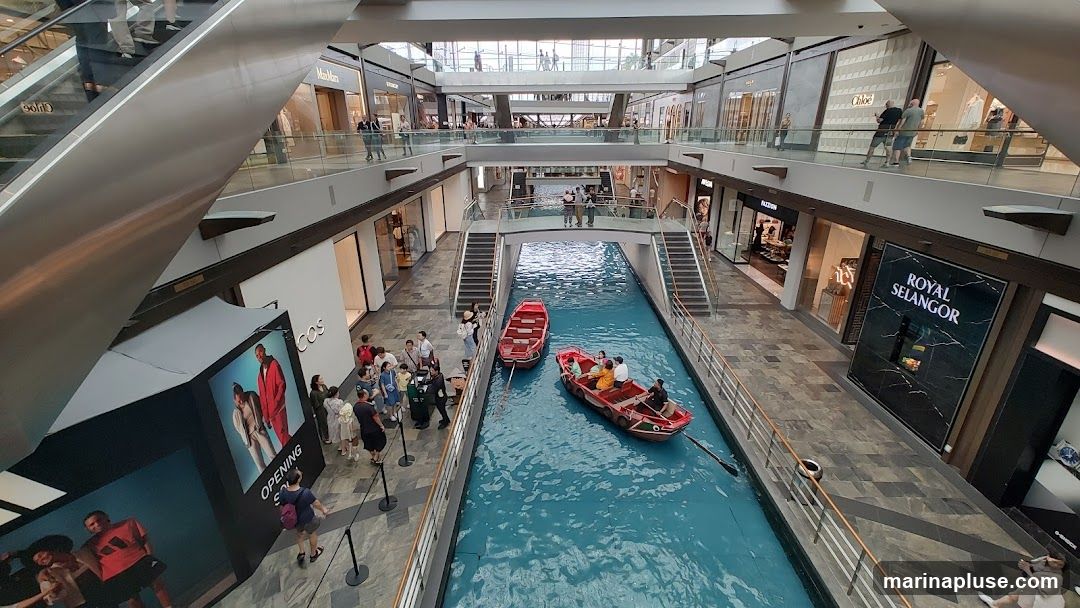 An interior view of a multi-level shopping mall with a canal running through the center. Red gondola-style boats are being rowed by people on the canal. Shops are visible on both sides of the canal on multiple levels, with escalators and walkways connecting them. A large advertisement for COS is on the left, and a sign for Royal Selangor is on the right.
