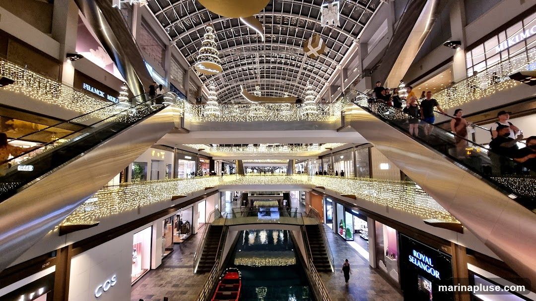 The image shows the interior of a multi-level shopping mall, decorated with numerous strings of warm white lights along the railings and ceilings. Escalators are visible, with people using them to move between floors. Shops with illuminated storefronts are arranged along the walkways. In the center, a canal with a boat is visible, reflecting the lights. Decorative elements, possibly for a holiday season, hang from the ceiling, including Christmas trees and abstract shapes. Brands like Franck Muller, COS, and Royal Selangor are visible on storefronts.