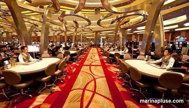 A wide shot of a bustling casino floor with numerous gaming tables and slot machines. People are seated at the tables, some playing cards, while others stand around. The room is elaborately decorated with ornate pillars, curved ceiling structures, and a red carpet with a swirling pattern running down the center aisle. The lighting is warm and inviting, creating a lively atmosphere.