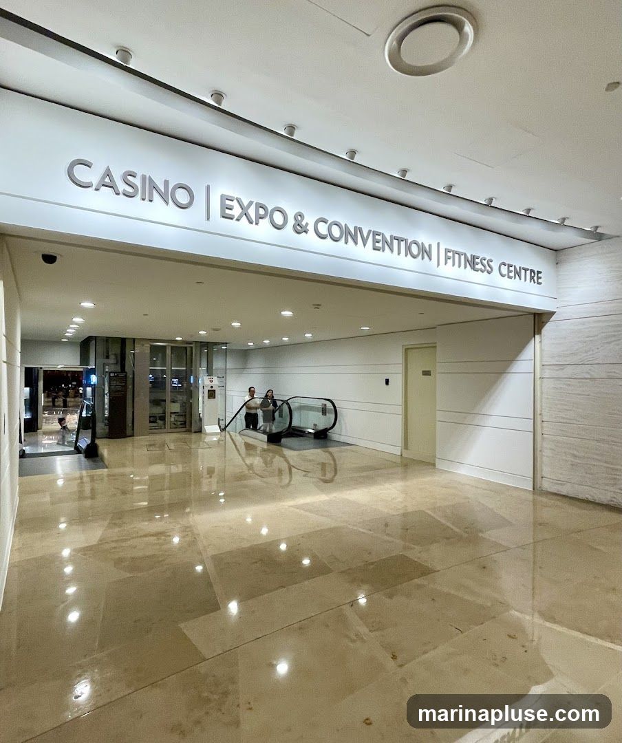 An interior view of a modern building lobby with polished marble floors. A large illuminated sign above reads "CASINO | EXPO & CONVENTION | FITNESS CENTRE". To the left, a glass-enclosed entrance leads to an area with bright lights. In the center, an escalator ascends, with two people standing near its base. A closed door is visible on the right wall.