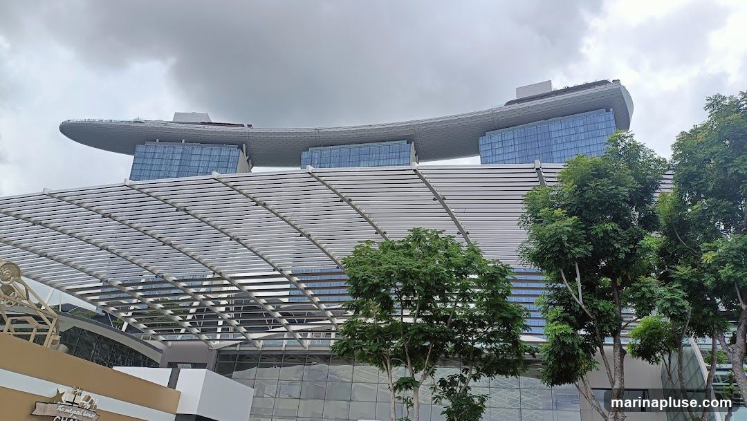 A view of the Marina Bay Sands hotel in Singapore, with its distinctive boat-shaped top structure. The foreground shows a modern architectural canopy and lush green trees. The sky is overcast with clouds.