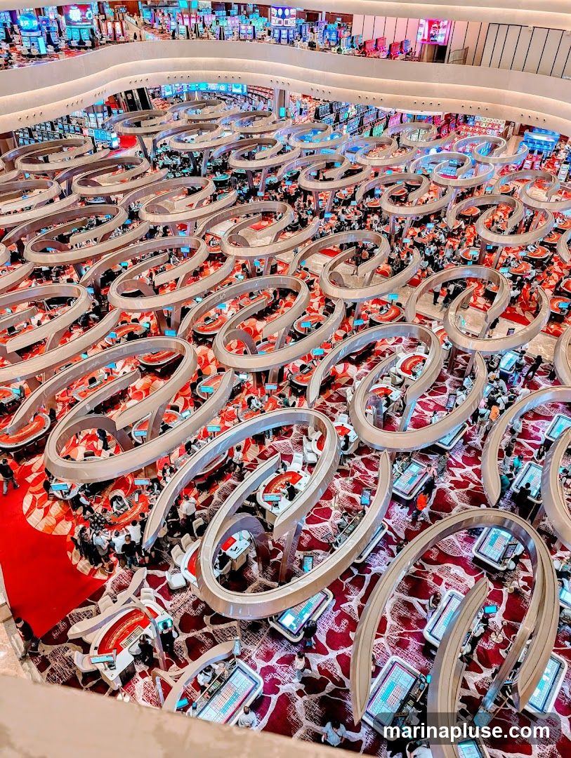 An aerial view of a bustling casino floor filled with numerous gaming tables and slot machines. People are gathered around the tables, engaged in various games. The floor is carpeted with a red and white patterned design, and the gaming tables have a modern, curved aesthetic with integrated screens. The overall impression is one of a vibrant and active entertainment venue.