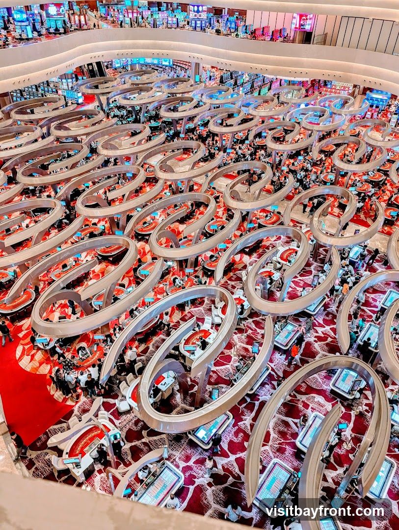 An aerial view of a bustling casino floor filled with numerous gaming tables and slot machines. People are gathered around the tables, engaged in various games. The floor is carpeted with a red and white patterned design, and the gaming tables have a modern, curved aesthetic with integrated screens. The overall impression is one of a vibrant and active entertainment venue.