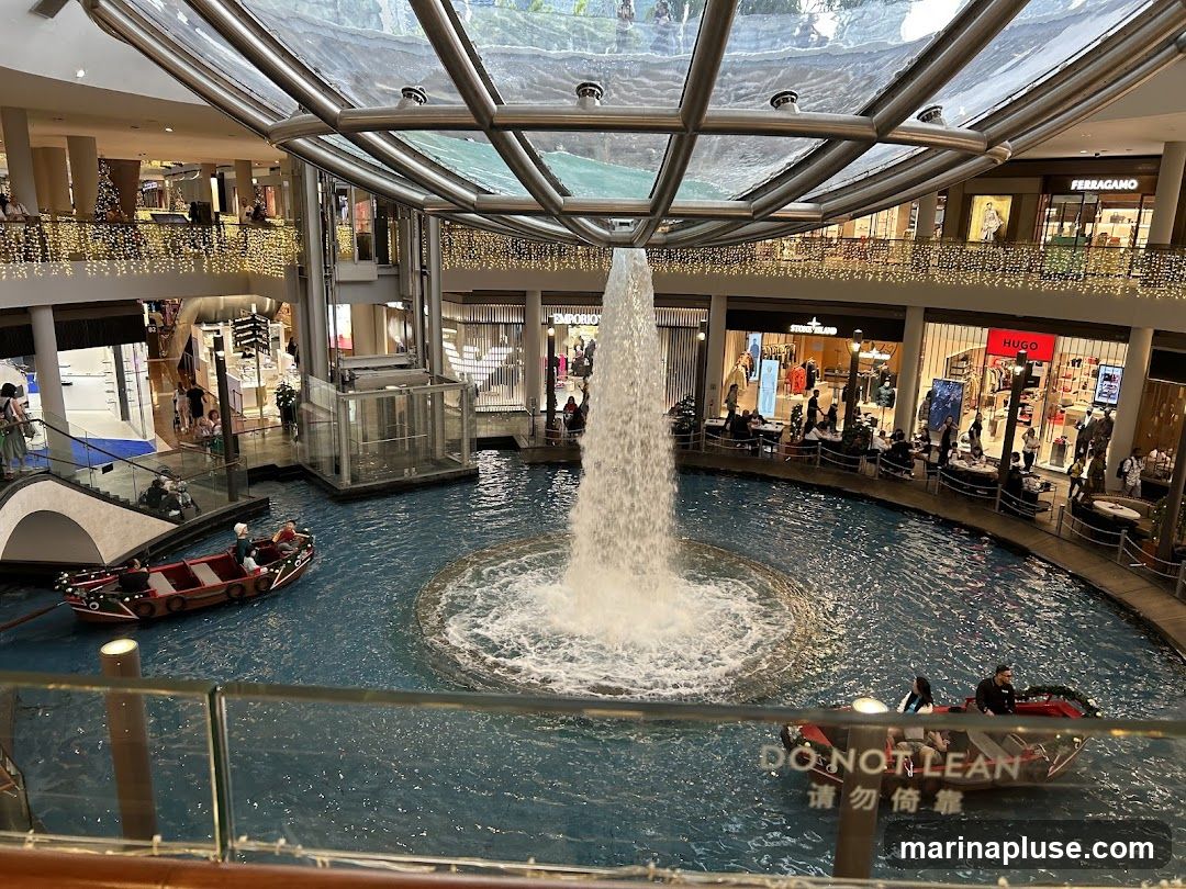 An interior view of a large, multi-level shopping mall featuring a central water feature with a large fountain. Small boats navigate the water, carrying passengers. Shops with visible signage like Ferragamo, Stone Island, and Hugo are present on the upper levels. Decorative lights adorn the balconies.
